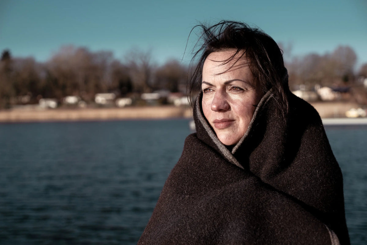 Eine Frau am Meer ist in unsere Decke "Airde" aus Walkloden gekuschelt und guckt in die Sonne.